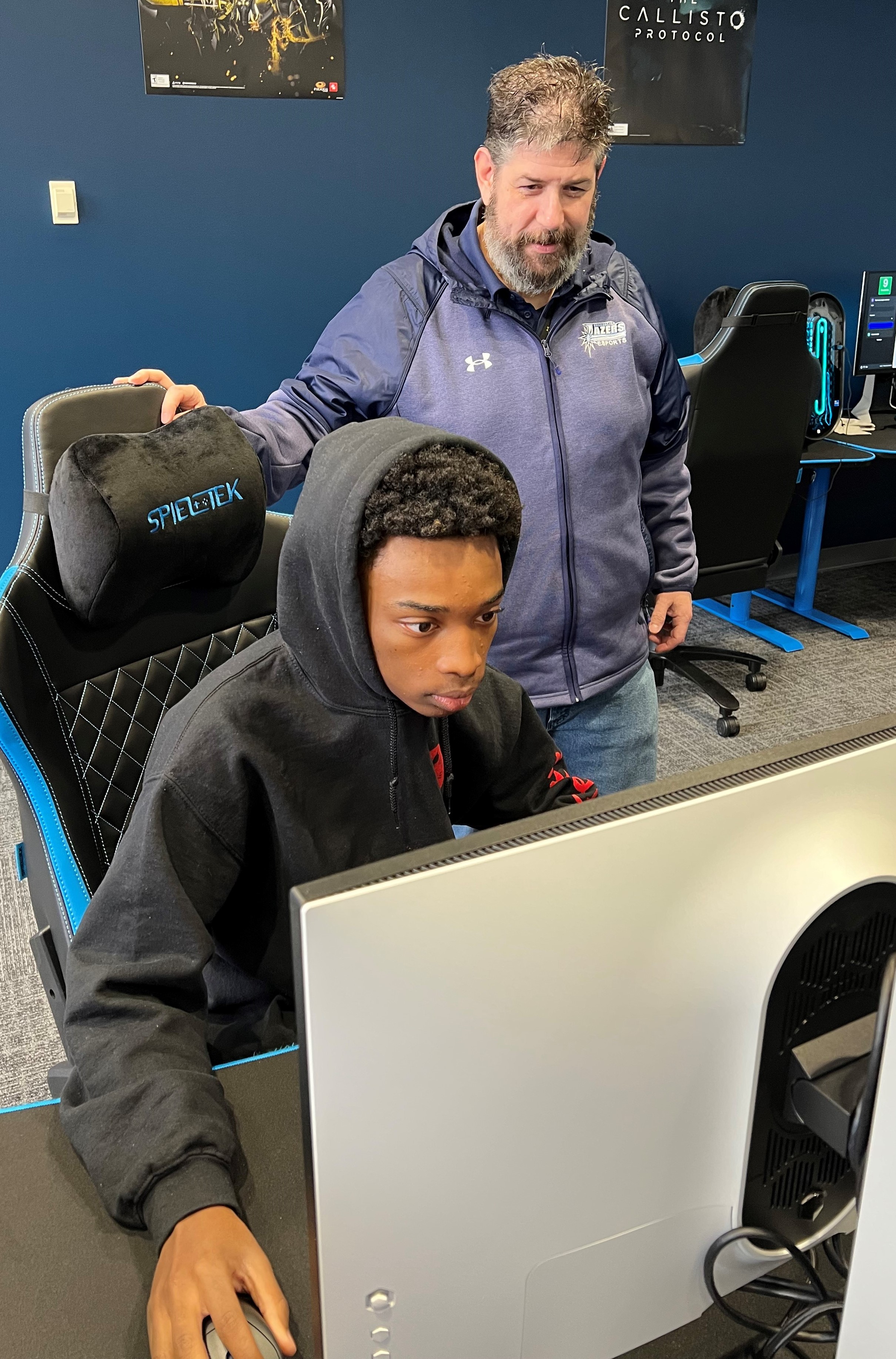 Esports Head Coach Keith Edwards (standing) watches student-athlete Dion Spaights (seated).