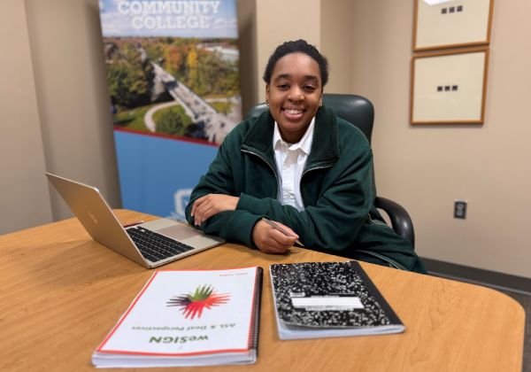 Student sitting at a table with a laptop and notebooks, Onondaga Community College banner in the background.