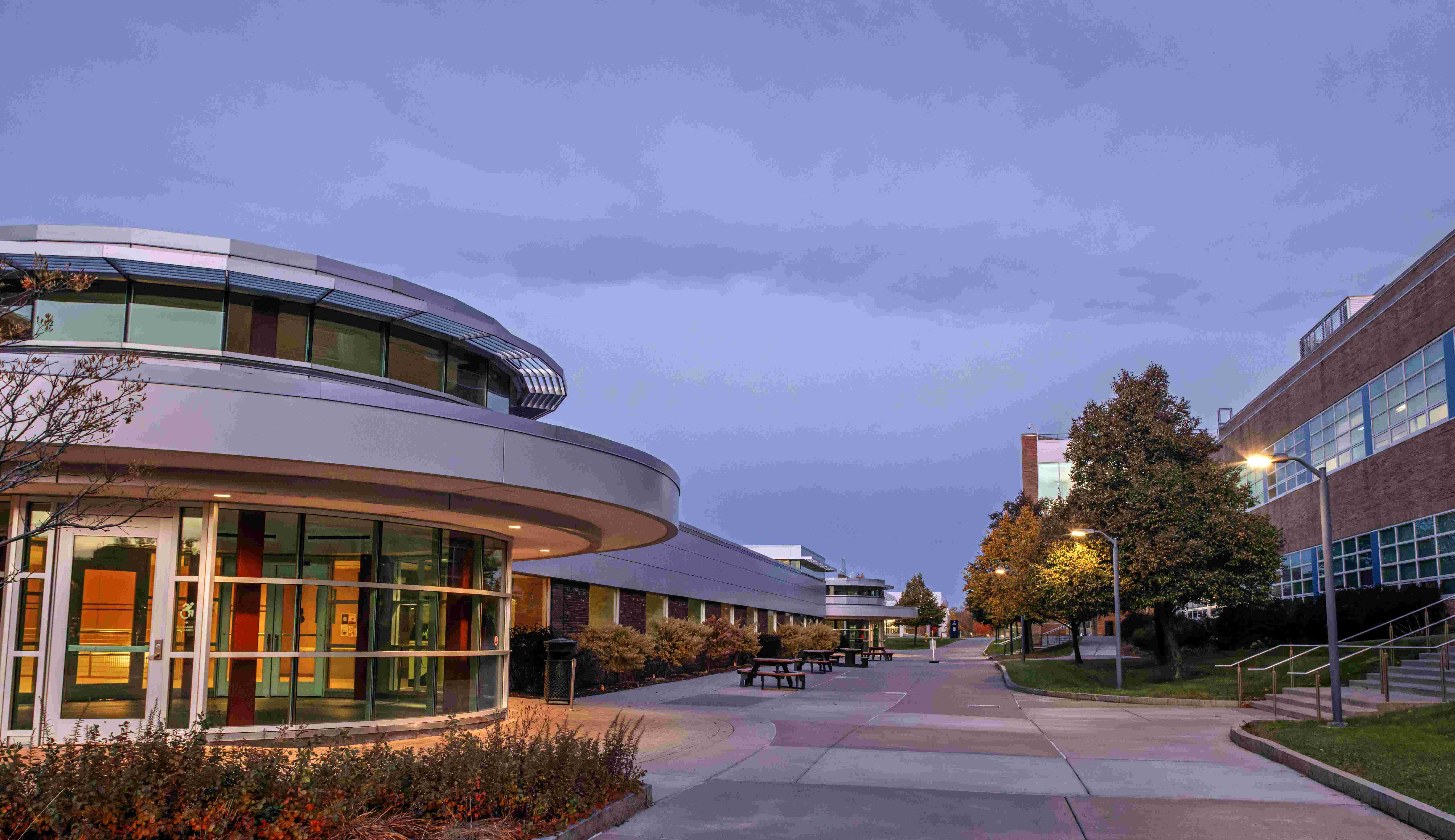 Modern campus buildings at dusk with pathway, benches, and streetlights.