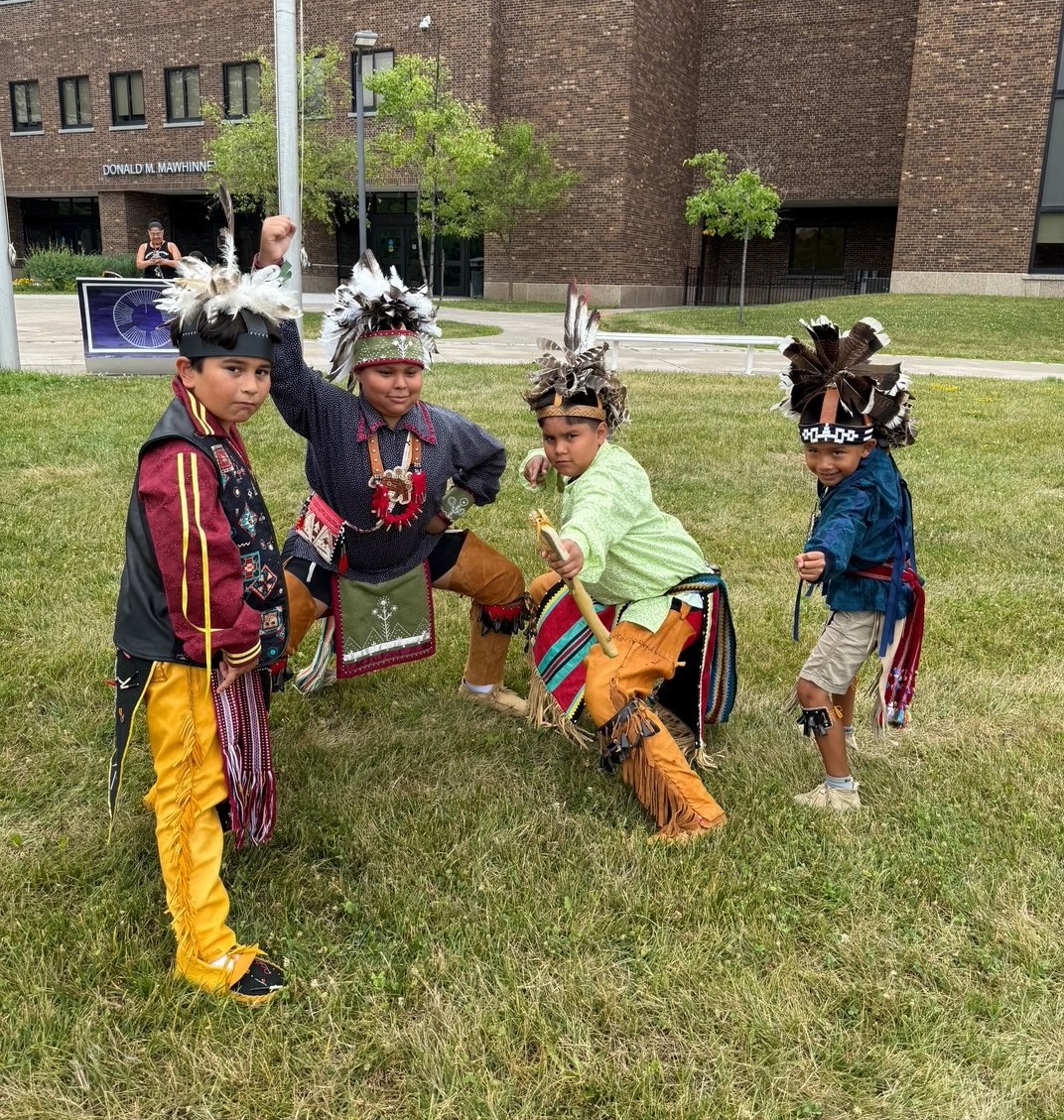 Four boys in traditional Native American attire posing outdoors on grass in front of a brick building.