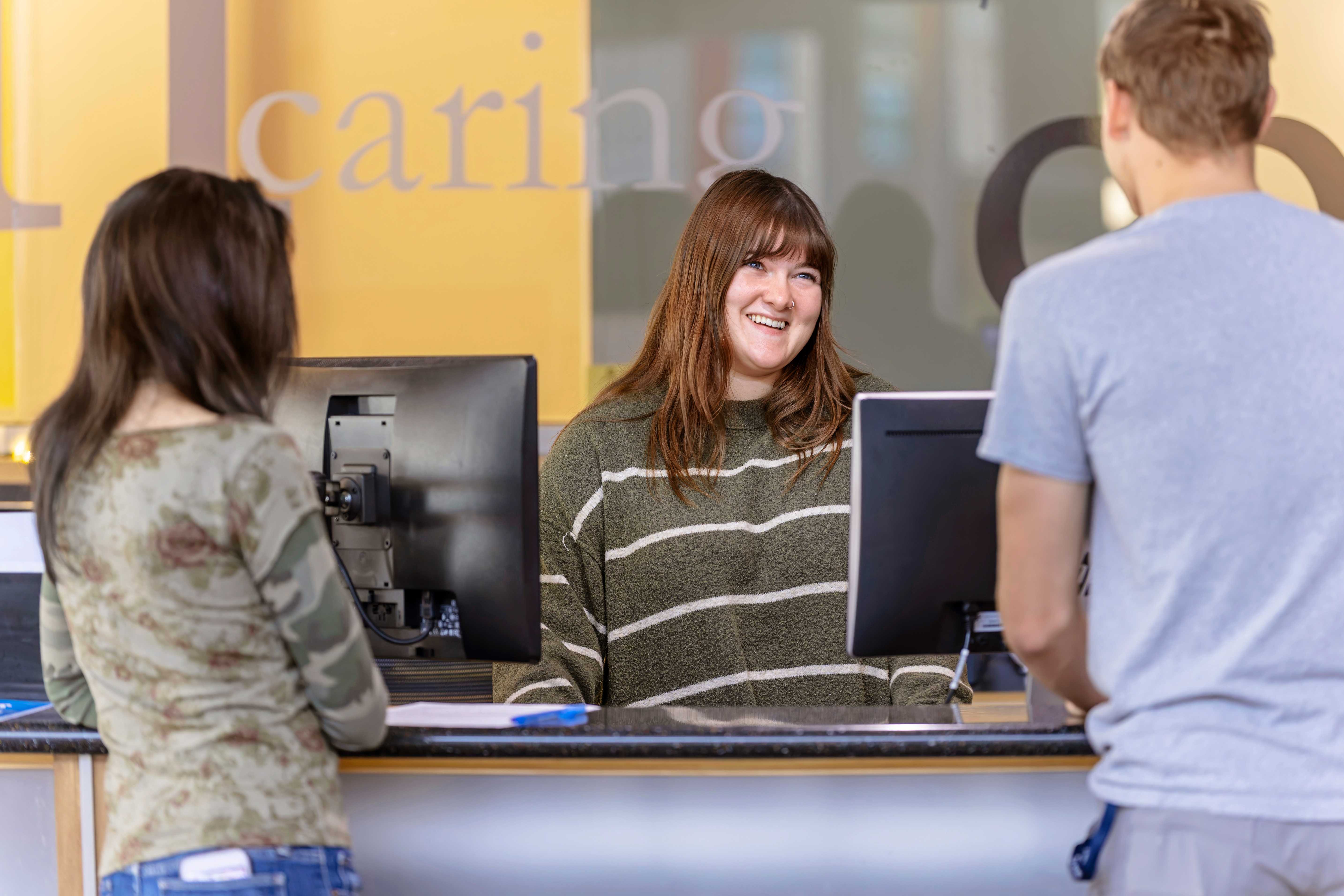 A smiling receptionist at a service counter assisting two people.