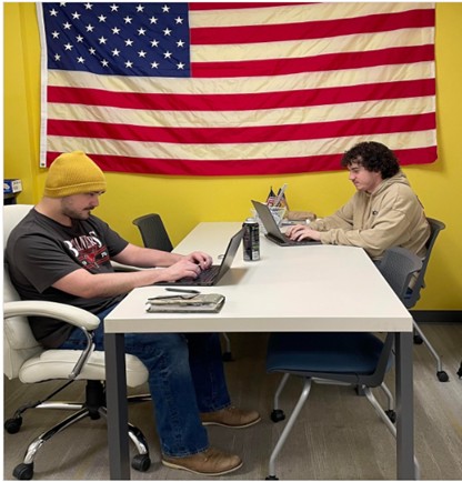 Two people sit across from each other at a table working on laptops in a yellow room, with a large American flag hanging on the wall behind them.