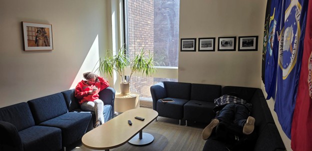 A lounge with dark sofas around a coffee table; one person in a red hoodie sits looking at a phone while another lies on a couch; sunlight streams through a tall window with plants nearby.