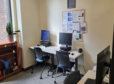 A small study room with two desktop computers on a white desk, office chairs, and a bulletin board on the wall; a window and potted plant sit to the left.