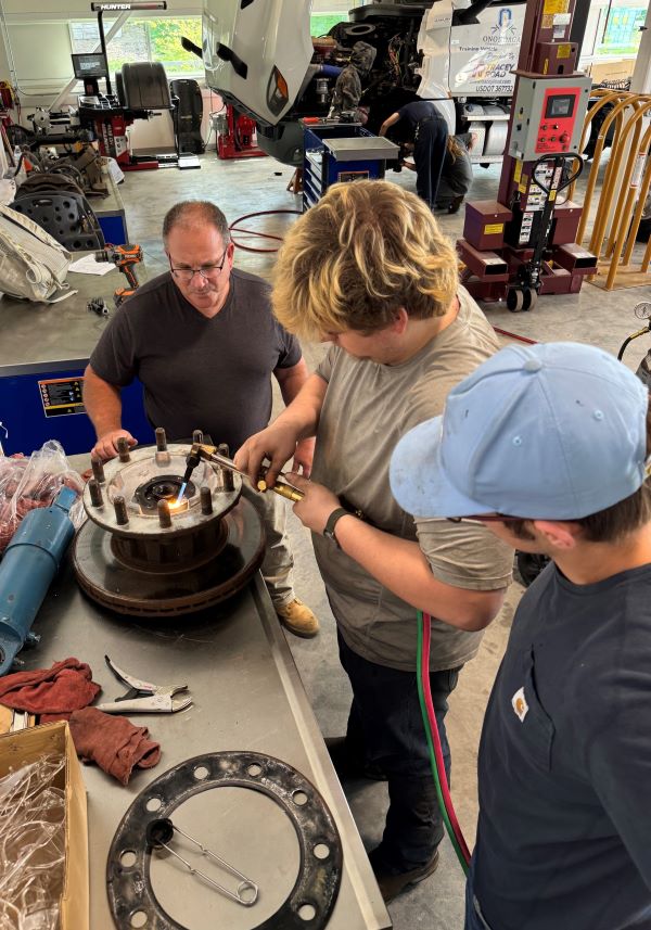 Professor Dave Lindsley (left) watches students Jackson Gwynn (center) and Donavan Boland (right) as they work to remove a broken bolt from a wheel.