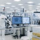 Semiconductor technician in a cleanroom wearing a full white protective suit and gloves, examining samples under a microscope at a workstation with computer monitors and fabrication equipment in the background.