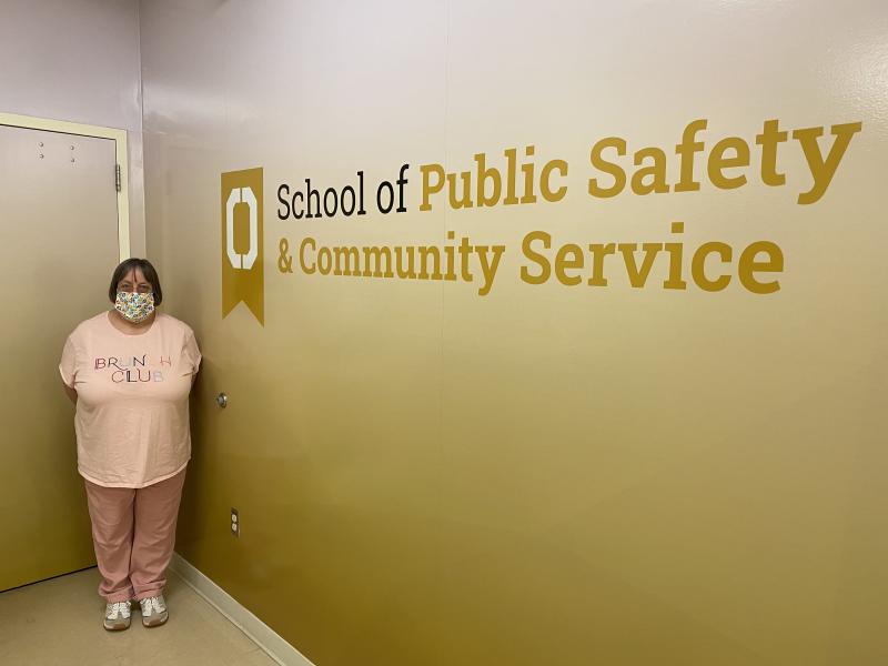 Woman of 53 years in age in a pink t-shirt that says brunch cluib stands in front of a yellow sign that says School of Public Safety and Community Service!