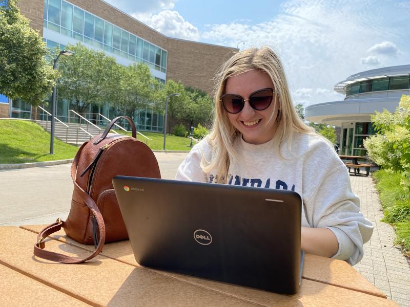 Woman with a Dell Chromebook and her tan bag sitting in the foreground of a building with glass windows. Blue skies can be seen in the background!