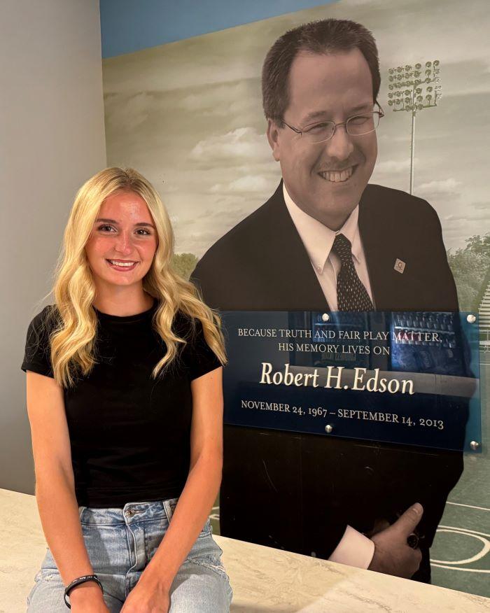 Ady Olmstead is the 2026-2027 recipient of the Robert H. Edson Memorial Scholarship. She's pictured in front of a wall honoring his legacy in the SRC Arena. Olmstead is a leader on the OCC Women's Soccer team and a Health Sciences major who plans to transfer to SUNY Upstate Medical University.