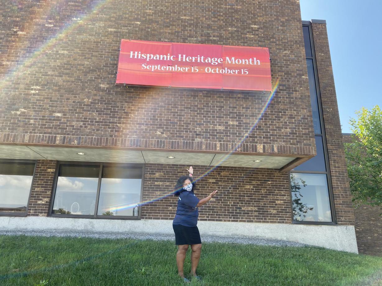 Woman with a mask standing in front of a sign that reads "Celebrating Latinx Heritage Month September 15 - October 15"