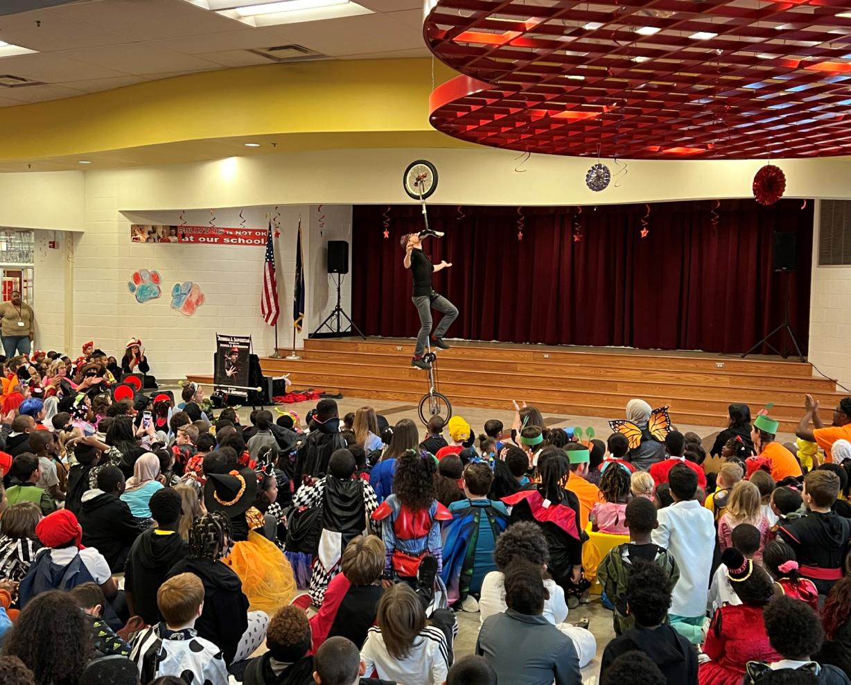 CirqOvation's Joshua J. Superstar rides a unicycle while holding another unicycle in his mouth in front of a packed cafeteria at Syracuse's Dr. Weeks School. The performance was coordinated by students in OCC's Phi Theta Kappa Honor Society.