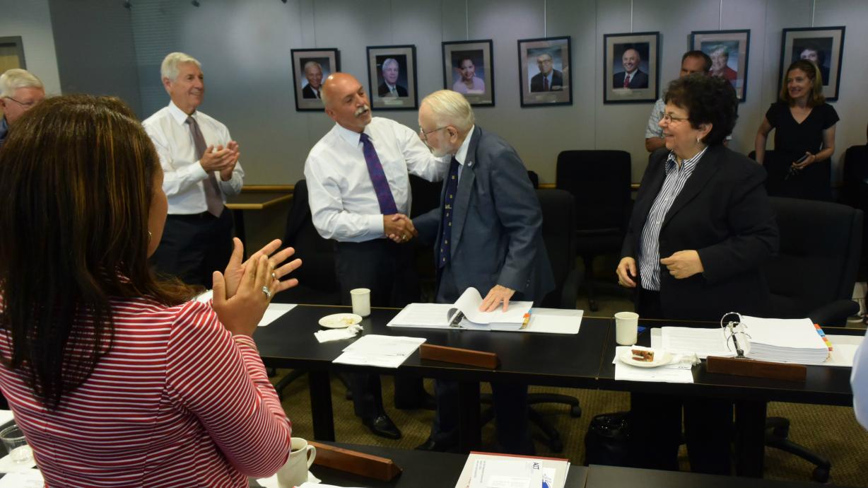 Donald Mawhinney (wearing blue blazer)  is congratulated by OCC Board of Trustees Chair Allen J. Naples and receives a standing ovation from fellow Trustees.